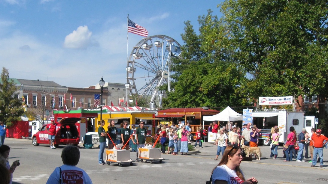 Ein klassisches Kleinstadtfest mit Riesenrad im Hintergrund, Imbisswagen und vielen Besuchern. Eine amerikanische Flagge ist gut sichtbar, und der Schauplatz scheint ein von historischen Backsteingebäuden umgebener Stadtplatz zu sein. Das Bild fängt mehrere typische Elemente von Kleinstadtfeiern ein: Fahrgeschäfte, Essensverkäufer und das Zusammenkommen der Gemeinschaft in einer charmanten Innenstadtkulisse.